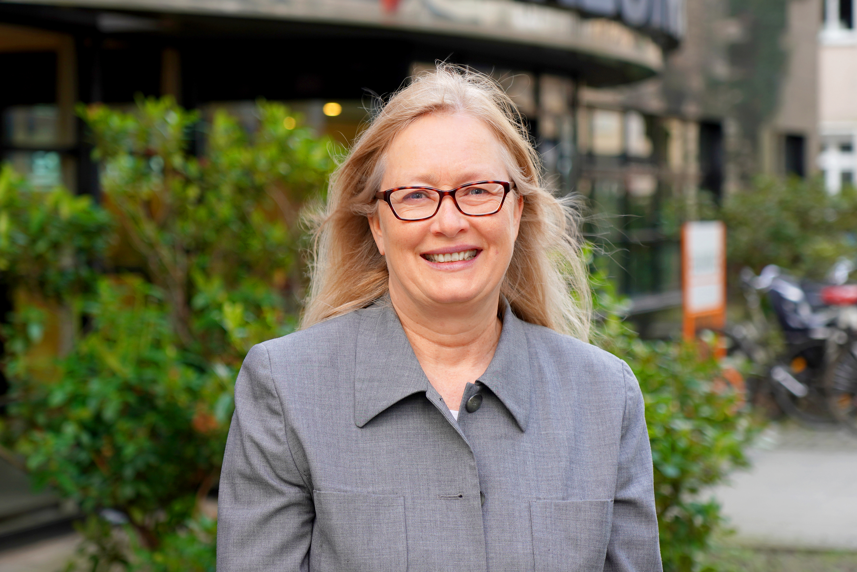 Zu sehen ist eine Frau mit blonden haaren und einer schwarzen Brille. Zudem trägt sie einen grauen Blazer und im Hintergrund sind Sträucher zu sehen.