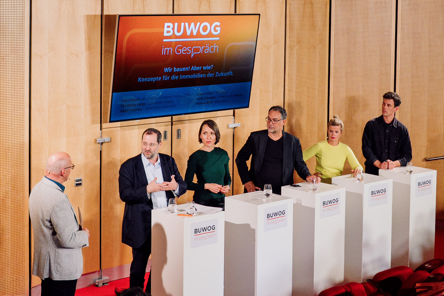 Zu sehen sind 6 Personen auf einer Bühne an Pulten stehend, die zu einem Thema diskutieren.