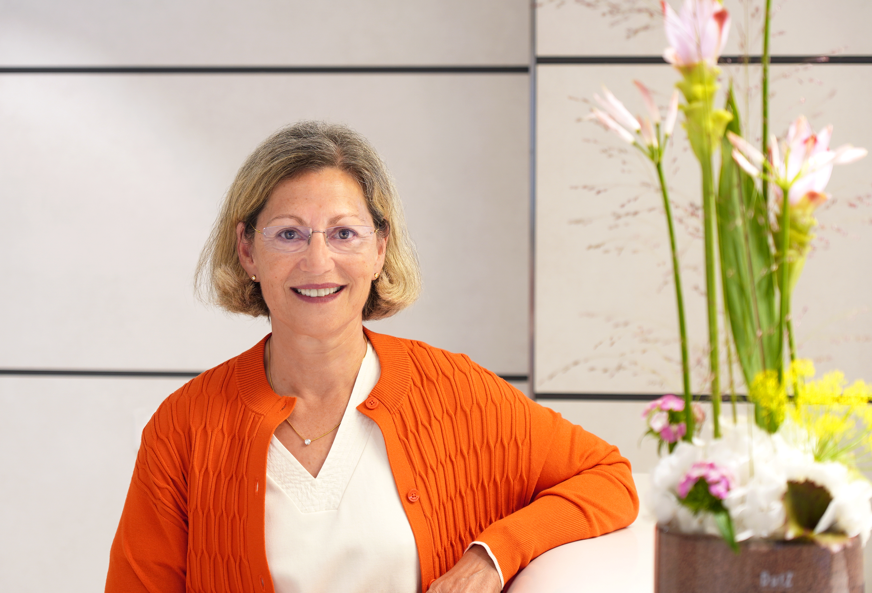 Zu sehen ist eine Frau mit blonden, kinnlangen Haaren in einer orangenen Strickjacke. Im Vordergrund stehen Blumen und der Hintergrund ist weiß. Sie lehnt ihren linken Arm auf einem Tresen an.