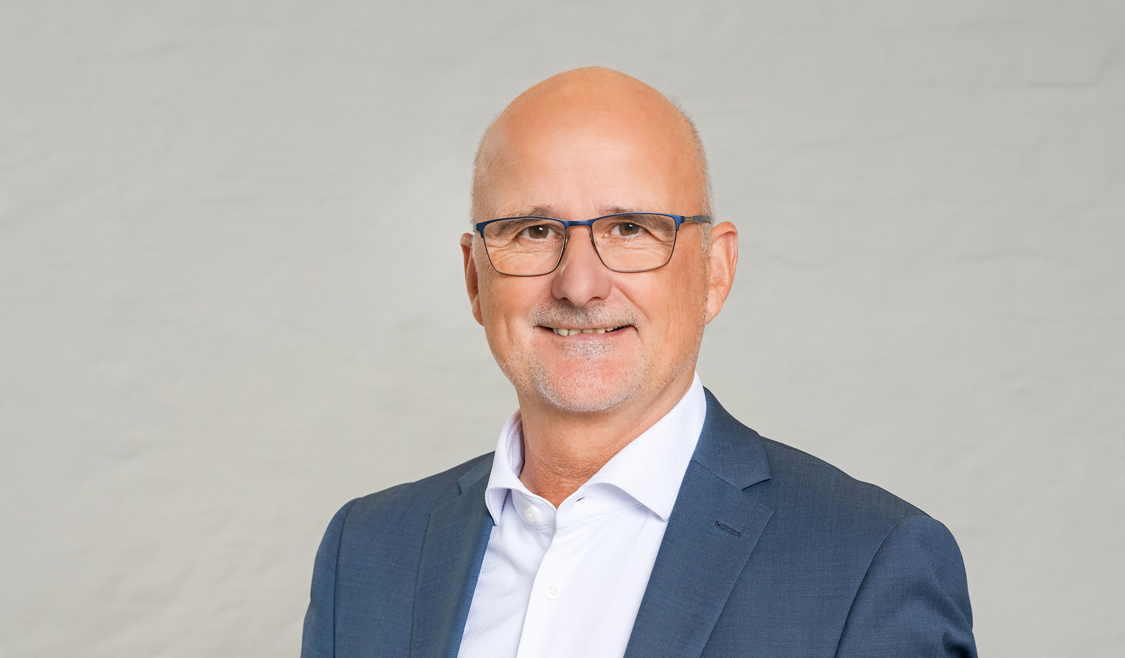 Zu sehen ist ein Mann in einem weißem Hemd und einem blauen Blazer. Er hat eine Glatze und eine blaue, große Brille.