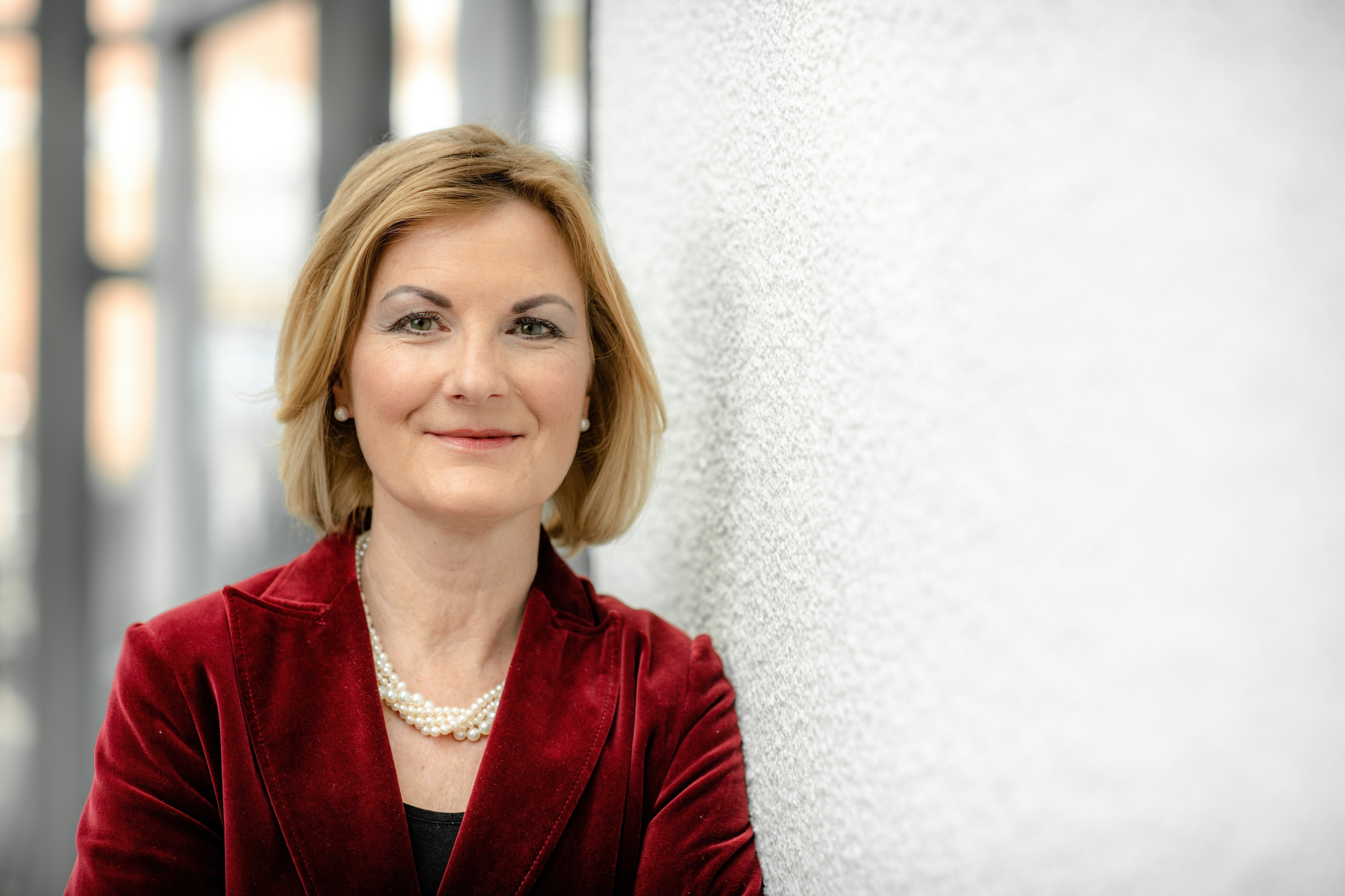 Zu sehen ist eine Frau, mit kinnlangen blonden Haaren, einem roten Samt-Blazer, einem schwarzen Oberteil und einer Perlenkette, anlehnend an einer Wand.